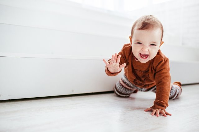 Un bebé gatea por el piso y saluda con la mano, riéndose