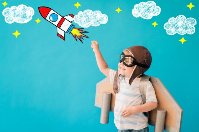 Un nene con gorro de aviador y un cohete de cartón juega a volar frente a una pared celeste