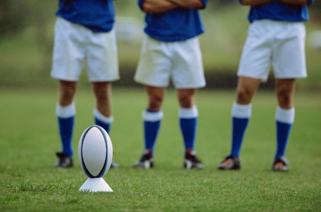 Una pelota de rugby sobre el tee frente a tres jugadores con medias azules